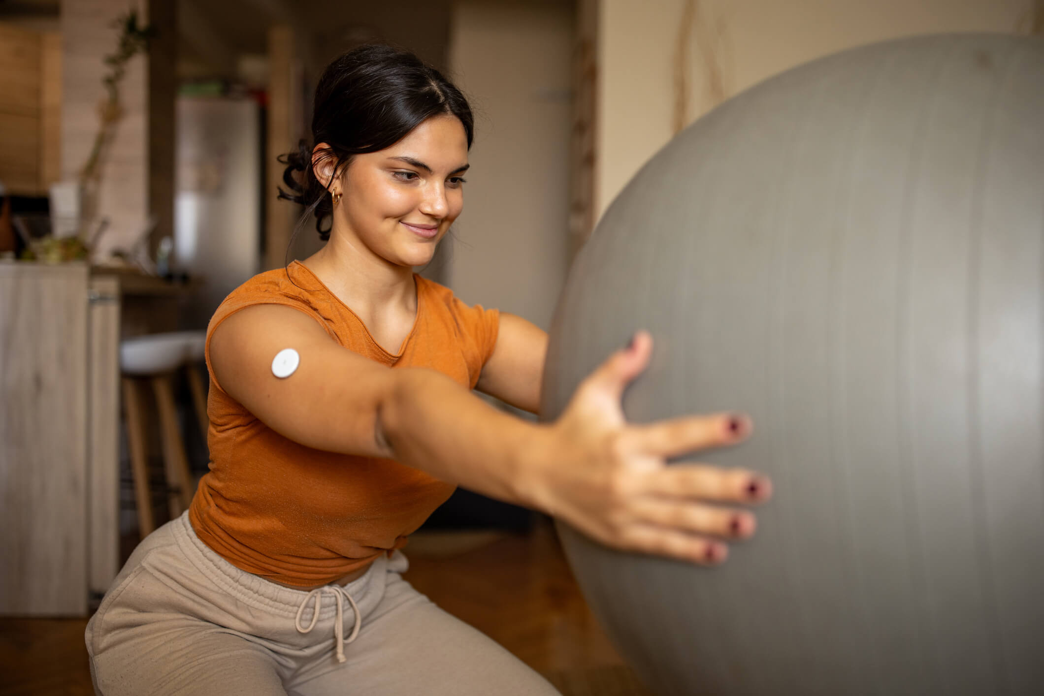 Young woman wearing a continuous glucose monitor squatting with a yoga ball held out in front of her.